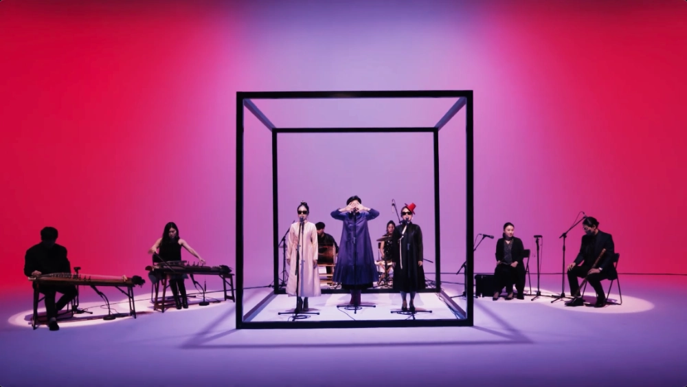 9 musicians in a soundstage lit with a soft purple red fill. the 3 vocalists are standing inside a large, black, cubic frame. 2 zither players are sitting to the right. 2 wind instrument players are to the left. 2 percussionists are sitting behind the cube.