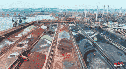 a birds-eye view of a mineral depot on a port. large piles of different black, grey, brown, and green ores are piled up next to water, cranes, and smokestacks