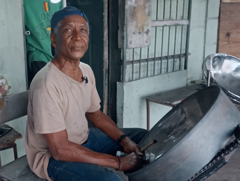 jimi phillip wearing a blue cap and shirt, sitting on a chair in front of an in-progress steelpan, tools in hand